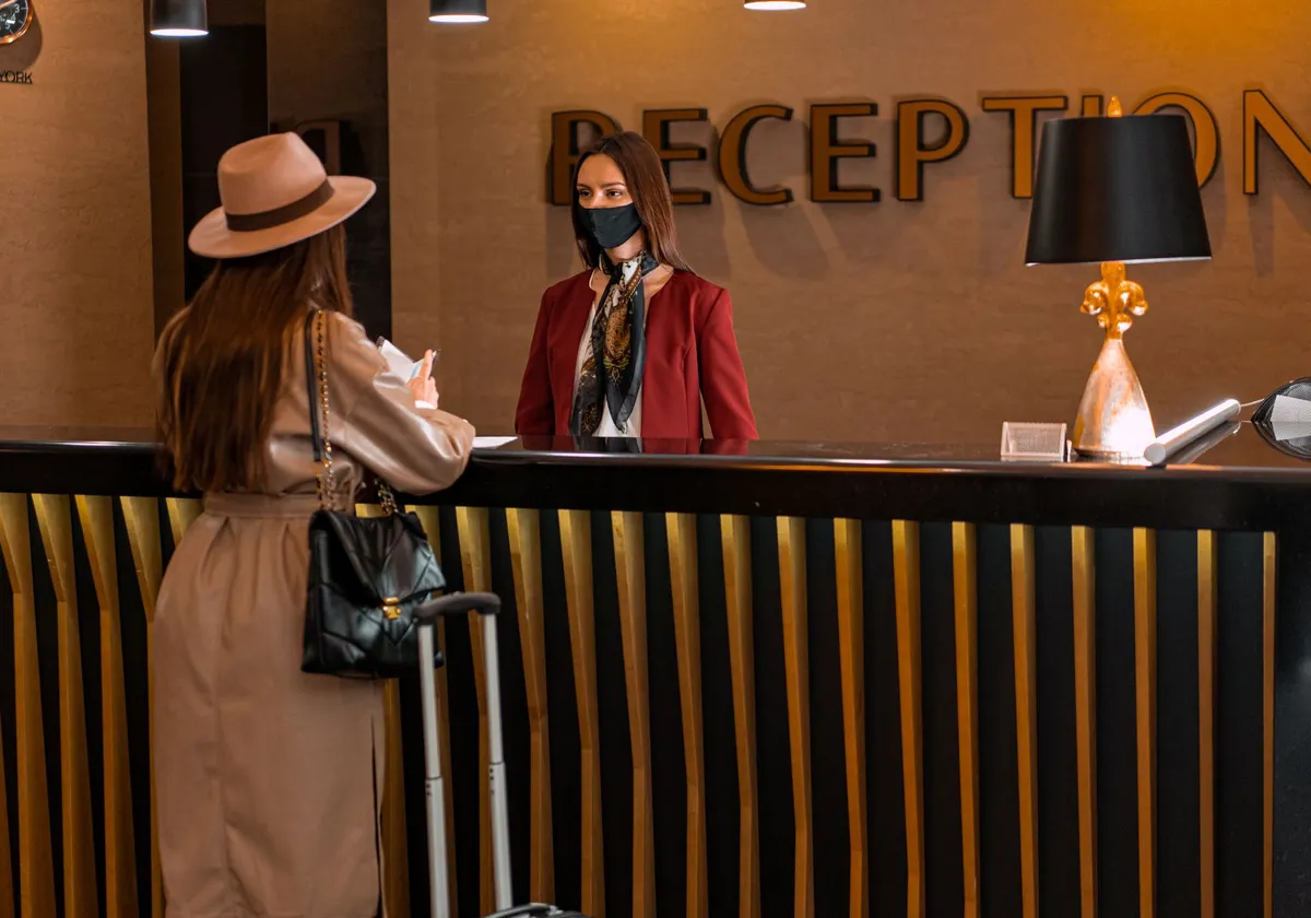 A woman speaks to a receptionist wearing a mask at a hotel reception.