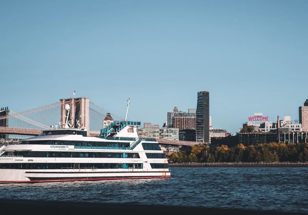 A cruise ship sails on the East River with the New York City skyline, including the Brooklyn Bridge, in view.
