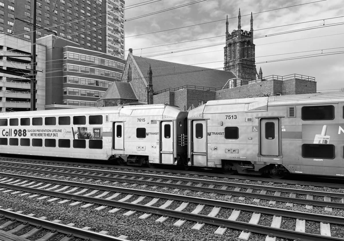 Black and white photo of NJ Transit train on urban tracks with city background.