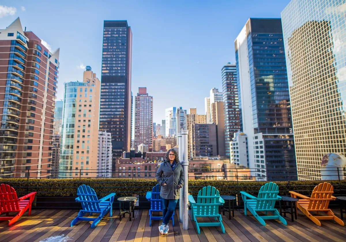 A woman stands on a rooftop terrace with colorful chairs against a city skyline.