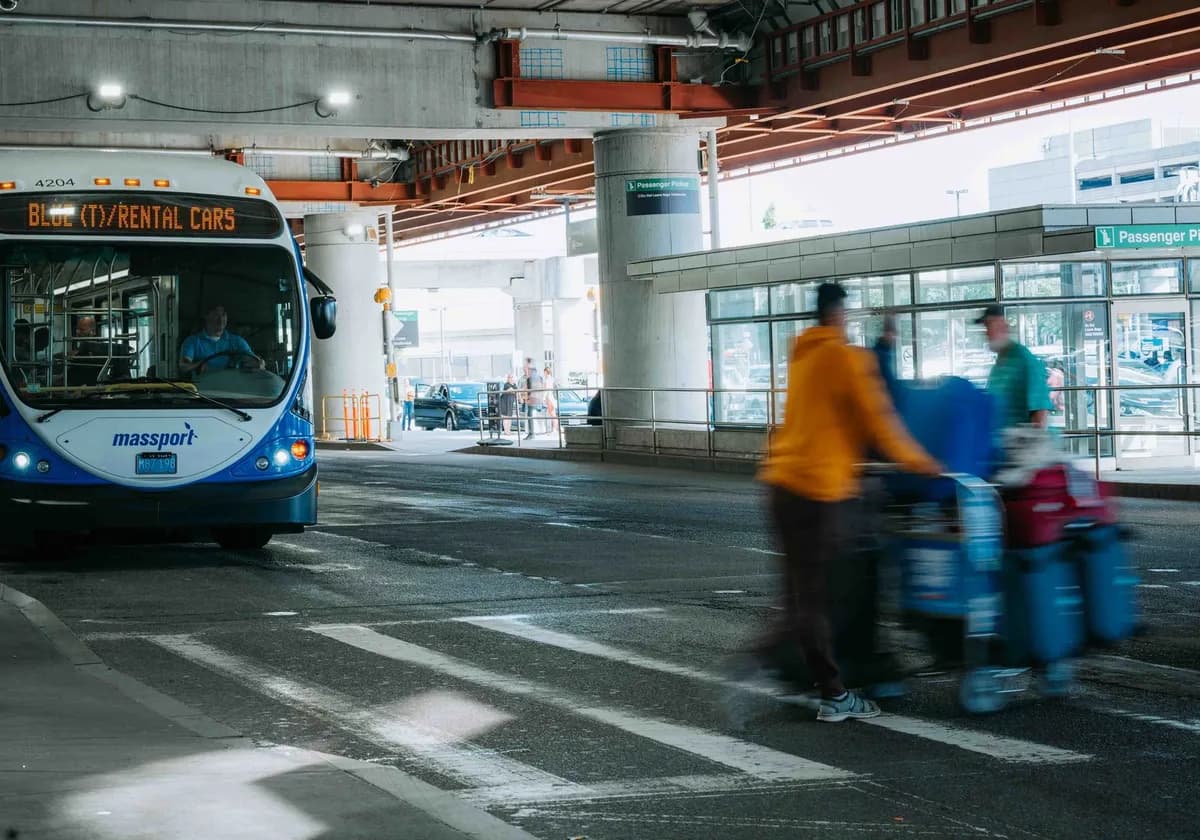 Busy airport scene showing travelers in motion and a rental car bus waiting under a covered terminal.