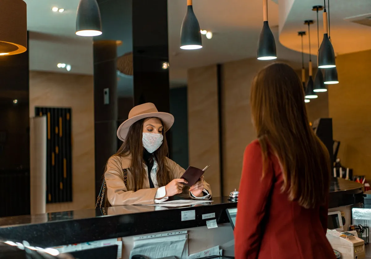A woman wearing a mask checks in at a hotel reception desk.