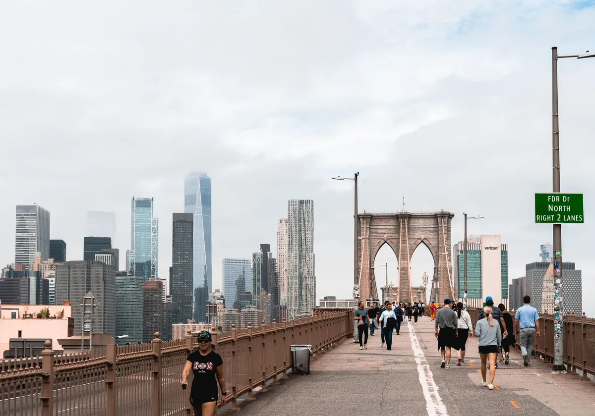 People walking on Brooklyn Bridge with Manhattan skyscrapers in the background under cloudy skies.